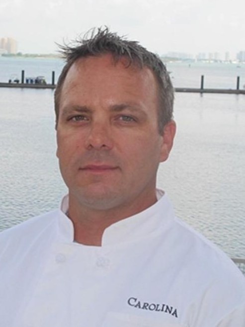 Andrew Siebert in whites on a dock, water and masts behind him
